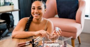 Smiling woman sitting at a table with acne-friendly makeup products including foundation, concealer, lipsticks, and brushes arranged neatly in a clean, soft lighting beauty setting.